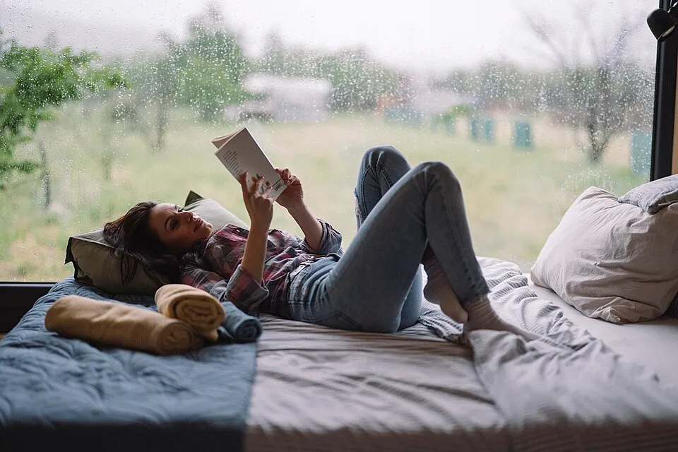 Woman relaxing with a book on a bed beside a rain-covered window