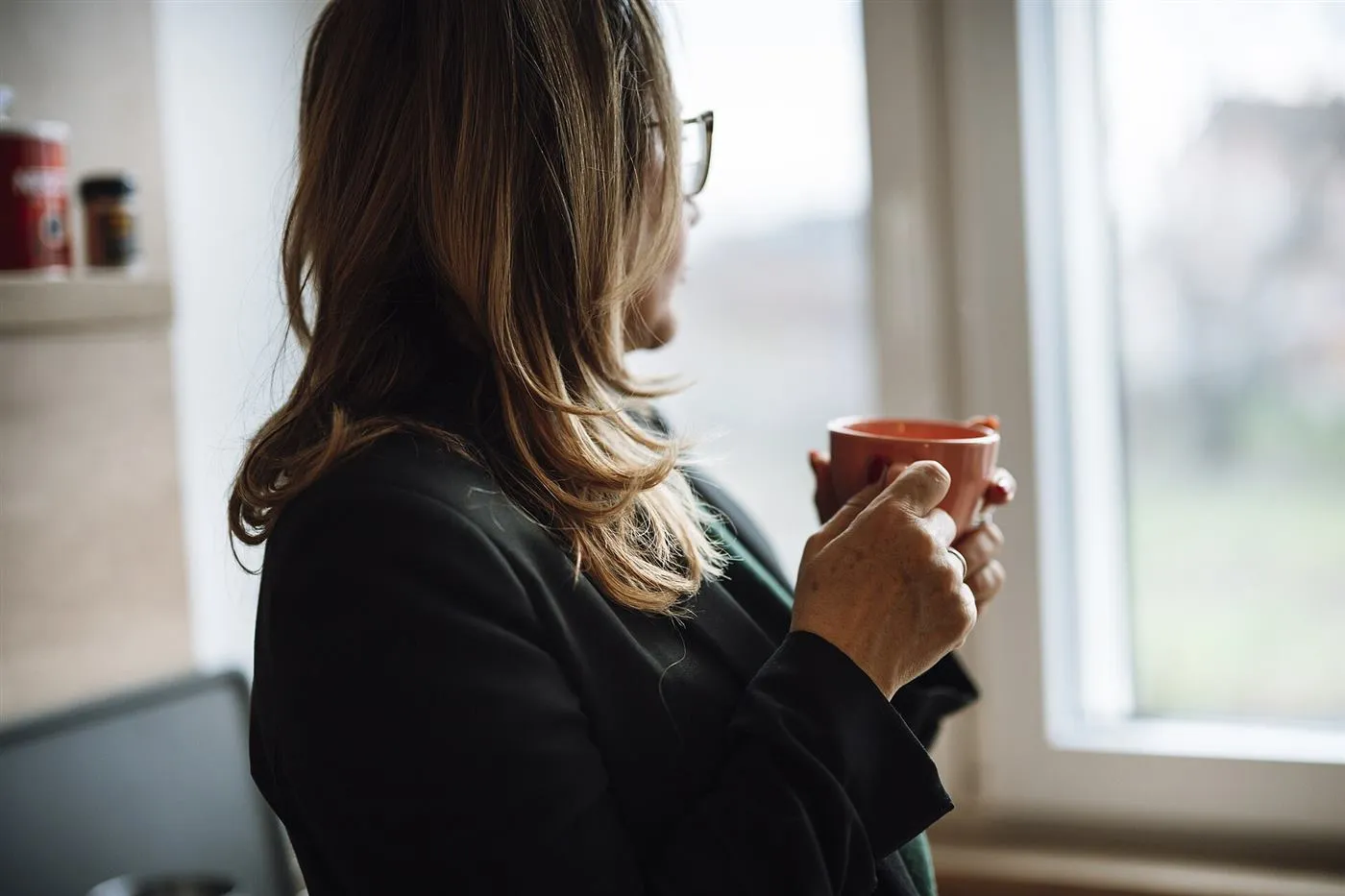 Woman holding a cup of coffee looking out her kitchen window in the morning
