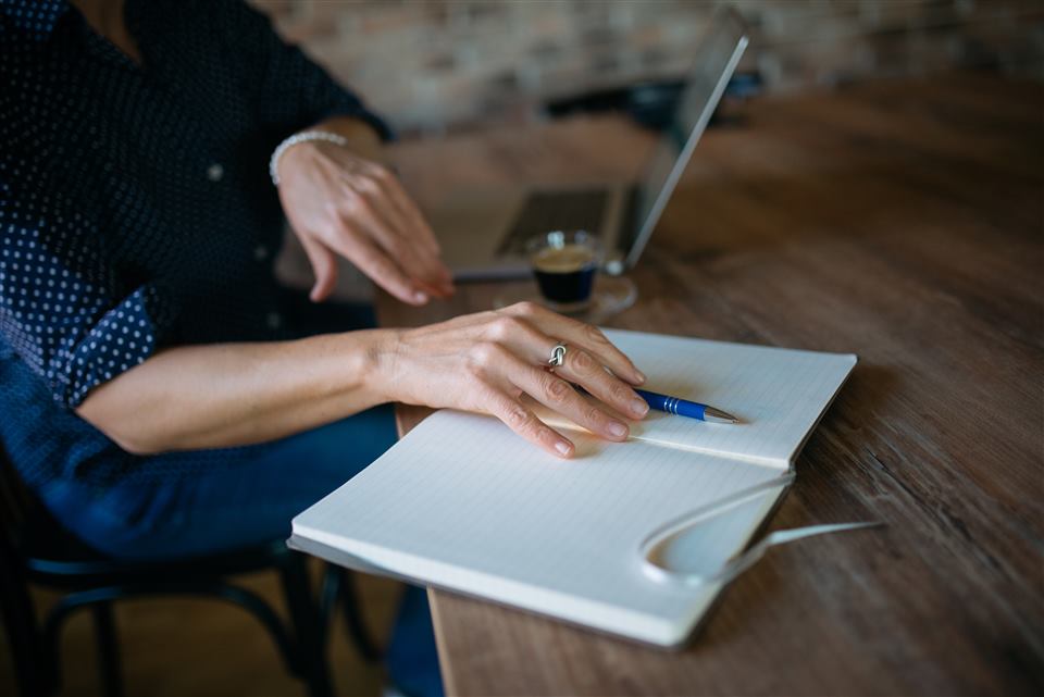 Close-up of a woman's hands with a pen resting on a blank open notebook, laptop and coffee in the background
