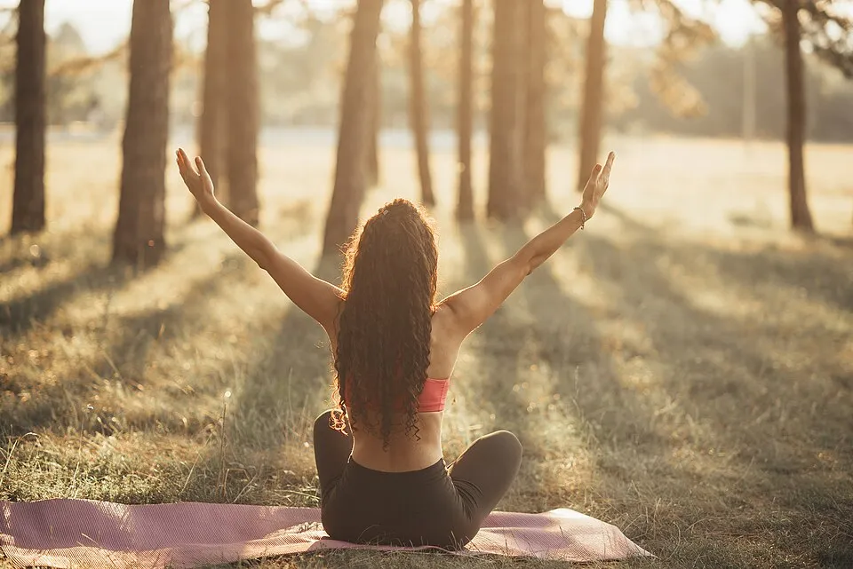 Woman sitting on a yoga mat in a sunlit pine grove at sunrise with her arms raised
